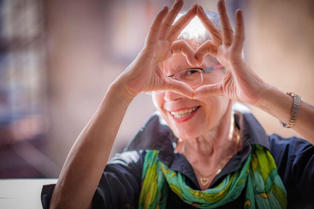 Woman with white hair in green scarf making a heart with her fingers around her eye